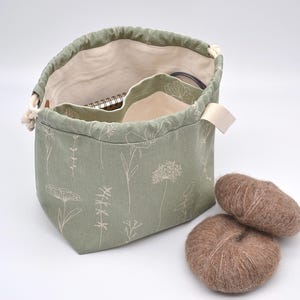 May include: Sage green drawstring project bag with a floral pattern, open to reveal a notebook and other items. Two brown yarn balls sit beside the bag. The bag has a cream-colored interior and a cream-colored strap.