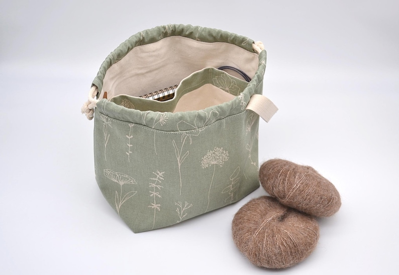 May include: Sage green drawstring project bag with a floral pattern, open to reveal a notebook and other items. Two brown yarn balls sit beside the bag. The bag has a cream-colored interior and a cream-colored strap.