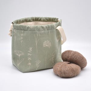 May include: A sage green drawstring bag with a floral pattern and two brown yarn balls. The bag has a cream-colored handle and a cream-colored drawstring. The yarn balls are fluffy and soft.