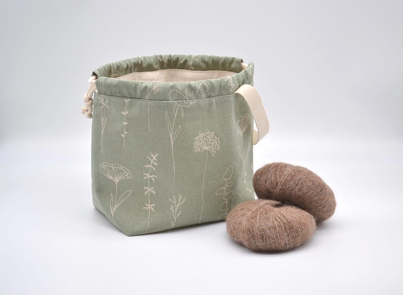 May include: A sage green drawstring bag with a floral pattern and two brown yarn balls. The bag has a cream-colored handle and a cream-colored drawstring. The yarn balls are fluffy and soft.