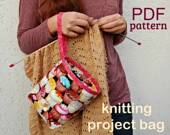 May include: A PDF pattern for a knitting project bag. The bag is made of fabric with a cupcake print and has a pink handle. The bag is being held by a person who is knitting a beige shawl.