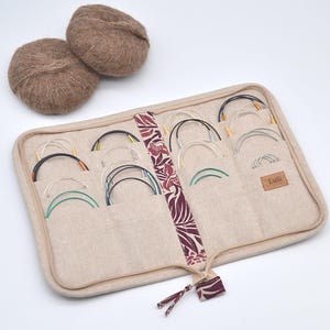 Puede incluir: Un estuche beige para agujas de tejer con m&uacute;ltiples bolsillos, que contiene varias agujas circulares con puntas de colores. Dos ovillos de lana marr&oacute;n se encuentran encima del estuche. El estuche tiene una franja floral decorativa y una peque&ntilde;a etiqueta de cuero que dice "Lusi".
