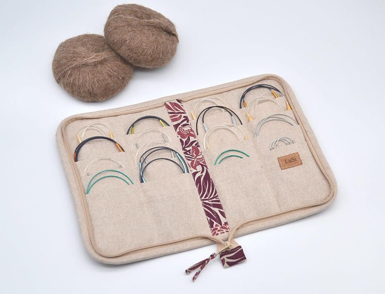 Puede incluir: Un estuche beige para agujas de tejer con m&uacute;ltiples bolsillos, que contiene varias agujas circulares con puntas de colores. Dos ovillos de lana marr&oacute;n se encuentran encima del estuche. El estuche tiene una franja floral decorativa y una peque&ntilde;a etiqueta de cuero que dice "Lusi".