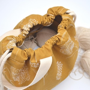 May include: Mustard yellow drawstring bag with a white floral pattern and cream-coloured handles. The bag is open, revealing white yarn inside. Two additional balls of yarn are next to the bag.
