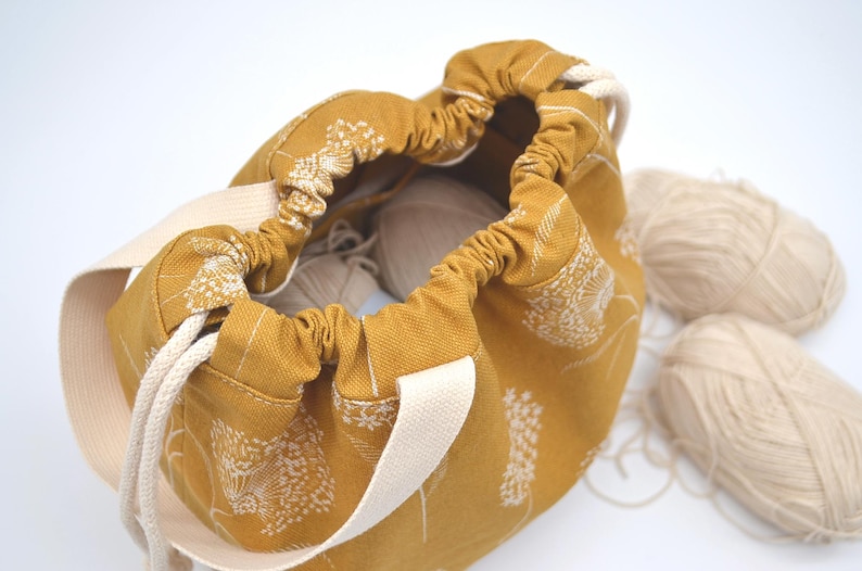 May include: Mustard yellow drawstring bag with a white floral pattern and cream-coloured handles. The bag is open, revealing white yarn inside. Two additional balls of yarn are next to the bag.