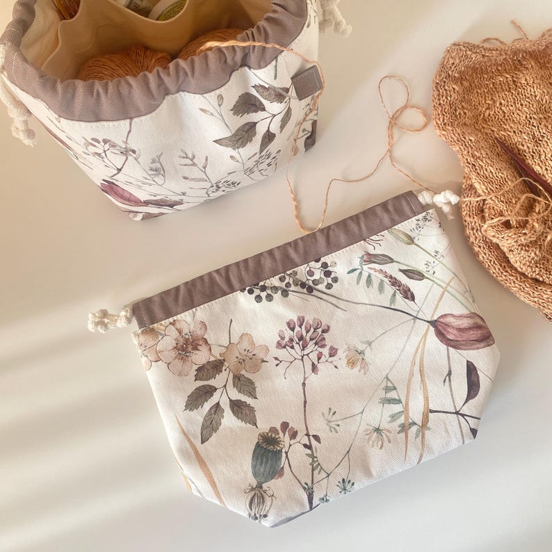 May include: Two drawstring project bags with floral patterns. The bags have a white background with a muted floral design in shades of brown, purple, and green. One bag is open, revealing yarn inside. The other bag is closed. A knitted item is also visible.