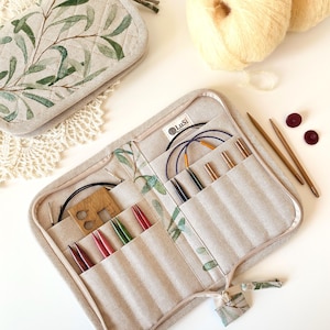 May include: A beige quilted fabric case with a green olive branch print, featuring a zipper closure and internal pockets holding various knitting needles, circular needle cords, and a wooden needle gauge tool, with a LuSi brand label inside.