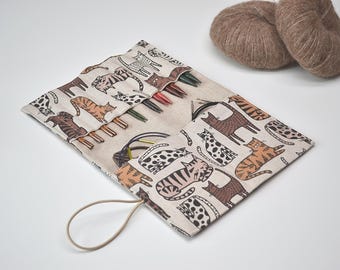 May include: A beige fabric knitting needle roll-up case featuring a repeating pattern of brown, orange, and black cats. The case contains multiple metal knitting needles and circular cables, secured with an attached elastic loop closure.