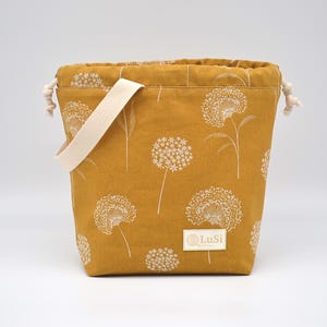 May include: Mustard yellow fabric bag featuring a white floral design and a cream handle. The bag has a drawstring closure and a small label with the text "LuSi".
