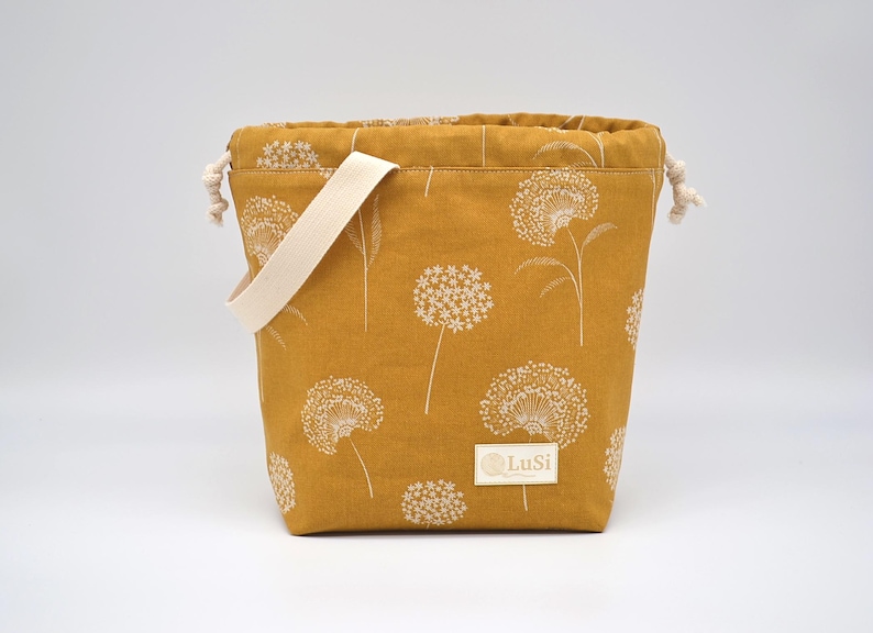May include: Mustard yellow fabric bag featuring a white floral design and a cream handle. The bag has a drawstring closure and a small label with the text "LuSi".