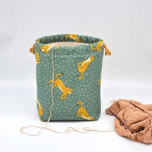 Op de afbeelding: Een teal stoffen garen opbergtas met een trekkoordsluiting. De tas heeft een herhalend patroon van gele cheeta's en kleine witte hartjes. Een bol garen en breinaalden liggen naast de tas.