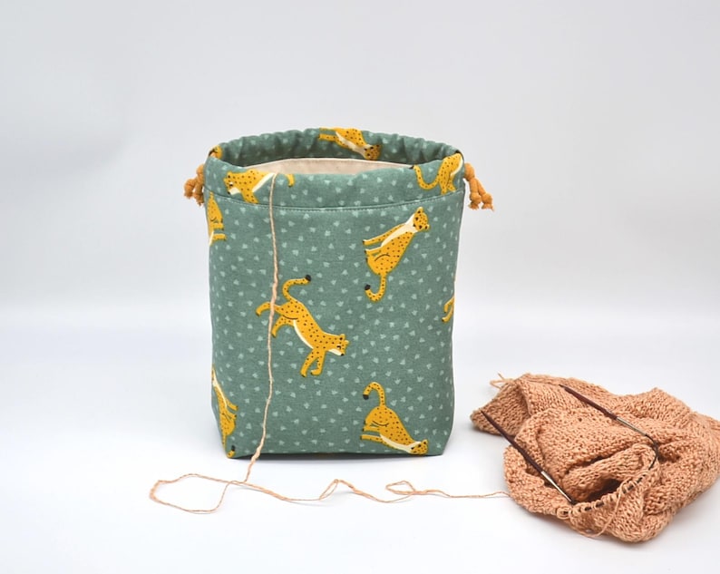 Op de afbeelding: Een teal stoffen garen opbergtas met een trekkoordsluiting. De tas heeft een herhalend patroon van gele cheeta's en kleine witte hartjes. Een bol garen en breinaalden liggen naast de tas.