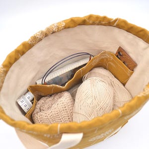 May include: Mustard yellow tote bag filled with knitting supplies. Inside are two balls of cream-coloured yarn, a small crocheted hat, a tape measure, and other accessories. The bag has a cream-coloured interior.