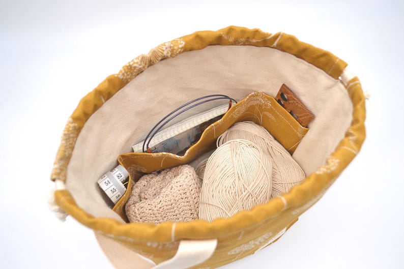 May include: Mustard yellow tote bag filled with knitting supplies. Inside are two balls of cream-coloured yarn, a small crocheted hat, a tape measure, and other accessories. The bag has a cream-coloured interior.