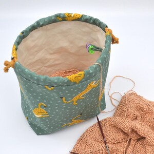 Op de afbeelding: Een turquoise projecttas met trekkoord en een herhalend patroon van gele cheeta's. De tas is open en onthult een lichtbruin interieur en een bol wol. Breinaalden en een gedeeltelijk gebreid stuk perzikkleurig garen zijn ook zichtbaar.