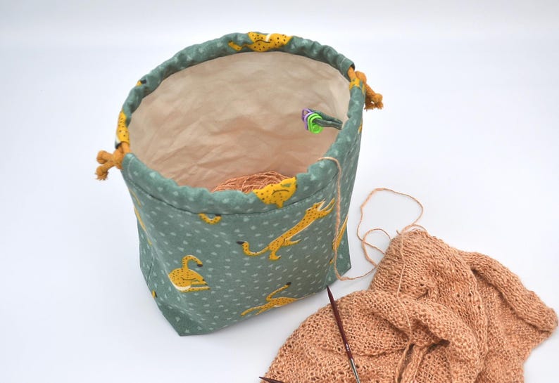Op de afbeelding: Een turquoise projecttas met trekkoord en een herhalend patroon van gele cheeta's. De tas is open en onthult een lichtbruin interieur en een bol wol. Breinaalden en een gedeeltelijk gebreid stuk perzikkleurig garen zijn ook zichtbaar.