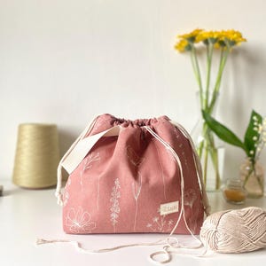 May include: A dusty rose drawstring bag featuring a white line-art floral pattern. The bag has a cream-colored fabric handle and a small rectangular label with the text LuSi sewn on the front. The closure is secured with thick cream-colored cords.