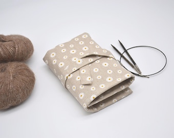 May include: A beige fabric knitting needle case with a white daisy print. The case is rolled up and tied with a beige ribbon. Two knitting needles and a black cord are next to the case. Two balls of brown yarn are on the left side of the image.