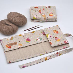 May include: A beige knitting needle case and accessories, decorated with colourful cupcakes and polka dots. Two balls of brown yarn and knitting needles are also present. The case has a label that reads "LuluSewing".