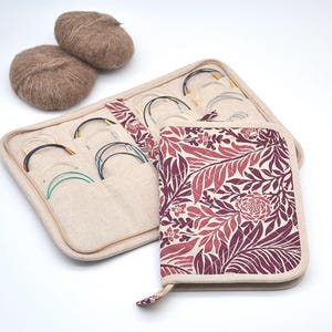 Puede incluir: Un estuche beige para agujas de tejer con un estampado floral en tonos burdeos y rosa, junto a un organizador beige con varias agujas de tejer circulares. Tambi&eacute;n hay dos ovillos de lana marr&oacute;n.