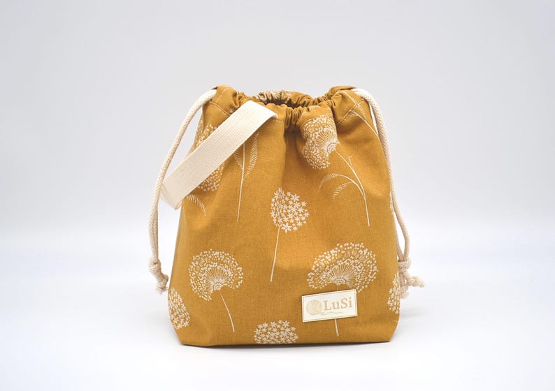 May include: Mustard-coloured drawstring bag with a white floral pattern. The bag has a cream-coloured strap and drawstring cords. A small rectangular label with the text "LuSi" is on the front.