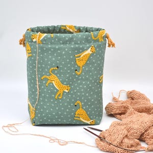 Op de afbeelding: Groene stoffen trekkoordtas met een patroon van gele cheeta's. De tas heeft een beige trekkoordsluiting. Een gebreide sjaal en breinaalden liggen ernaast. Ideaal voor het opbergen van knutselspullen.