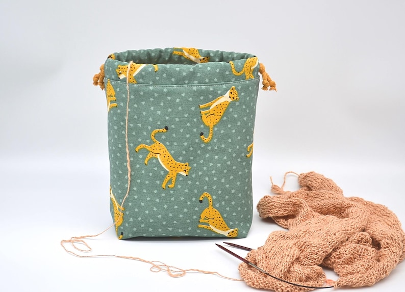 Op de afbeelding: Groene stoffen trekkoordtas met een patroon van gele cheeta's. De tas heeft een beige trekkoordsluiting. Een gebreide sjaal en breinaalden liggen ernaast. Ideaal voor het opbergen van knutselspullen.