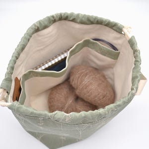 May include: A sage green drawstring bag with a floral pattern, containing two balls of brown yarn, a notebook, and knitting needles. The bag has a cream-colored interior with multiple pockets for organization.