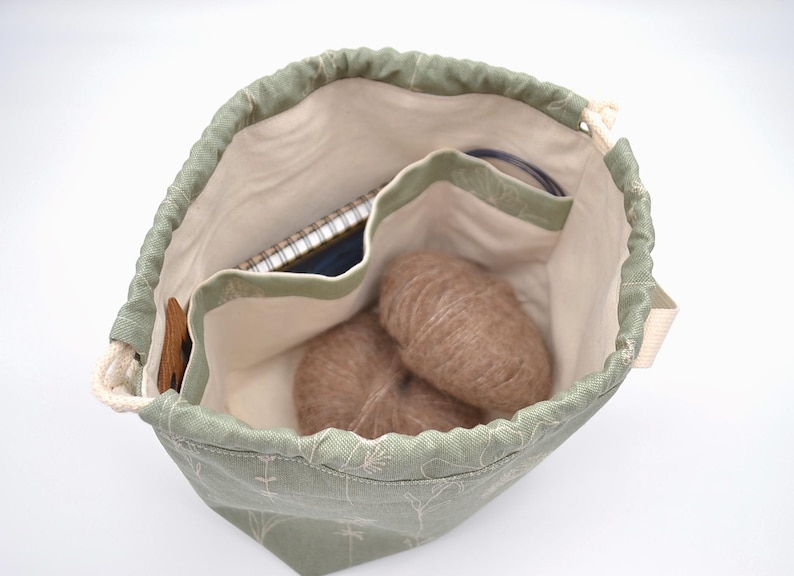 May include: A sage green drawstring bag with a floral pattern, containing two balls of brown yarn, a notebook, and knitting needles. The bag has a cream-colored interior with multiple pockets for organization.