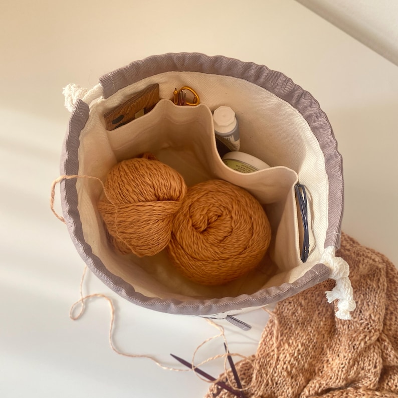 May include: A round, open-top knitting project bag with a gray rim and a drawstring closure. Inside are two balls of orange yarn, small tools, and a few other items. A partially knitted piece of fabric and knitting needles are visible.