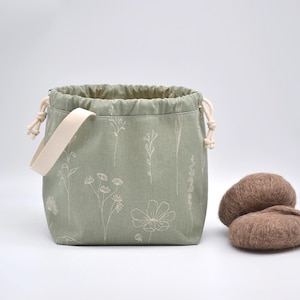 May include: Sage green drawstring bag with a cream-colored handle and floral embroidery. Two brown yarn balls sit beside the bag. The bag is designed for storing knitting or crochet supplies.