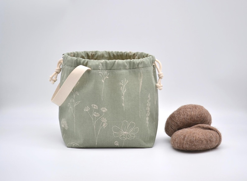 May include: Sage green drawstring bag with a cream-colored handle and floral embroidery. Two brown yarn balls sit beside the bag. The bag is designed for storing knitting or crochet supplies.