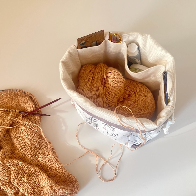 May include: A beige fabric knitting project bag with a floral pattern, holding two balls of orange yarn. The bag has several compartments containing scissors, a wooden tool, and other craft supplies. A partially completed knitted piece with needles is visible.
