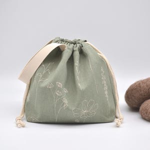 May include: Sage green drawstring bag with a cream-colored strap and drawstring. The bag is decorated with white floral embroidery. Two brown, oval-shaped objects are next to the bag.