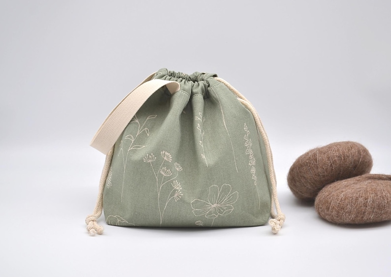 May include: Sage green drawstring bag with a cream-colored strap and drawstring. The bag is decorated with white floral embroidery. Two brown, oval-shaped objects are next to the bag.