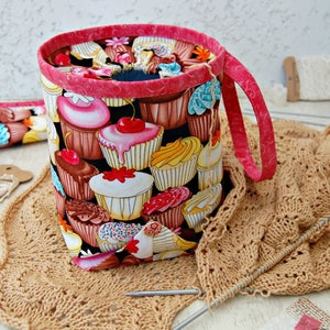 May include: A pink and black fabric yarn bowl with a cupcake pattern. The bowl has a handle and is sitting on a brown crocheted surface.