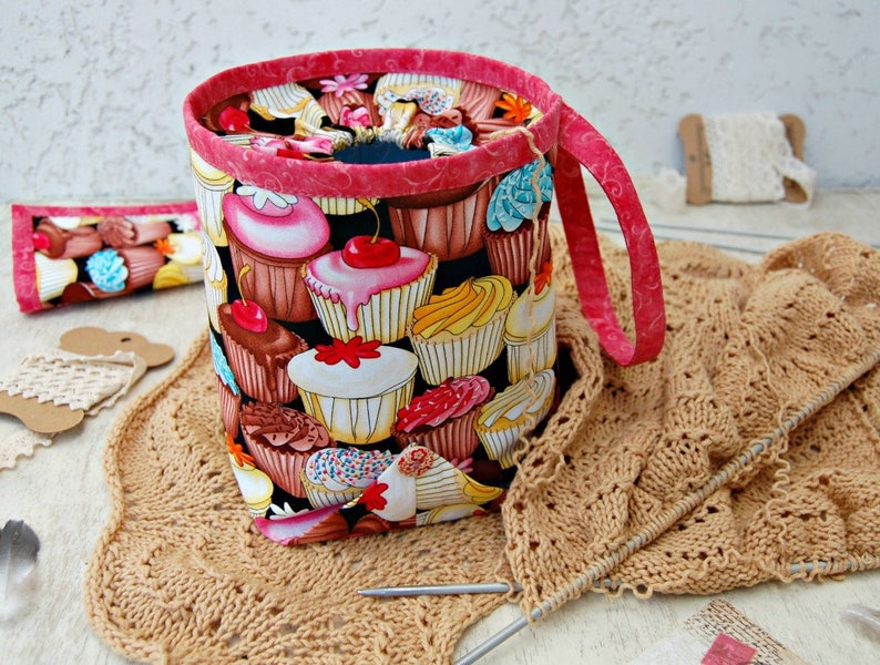 May include: A pink and black fabric yarn bowl with a cupcake pattern. The bowl has a handle and is sitting on a brown crocheted surface.