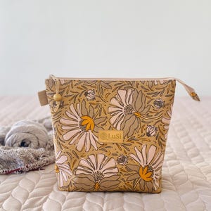 Op de afbeelding: Een tasje met rits met een mosterdgele achtergrond en een herhalend bloemenpatroon van witte en lichtroze bloemblaadjes met gele harten. Een rechthoekig beige label met de tekst LuSi is in het midden aan de voorzijde bevestigd. Een houten kraal aan de rits.