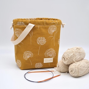 May include: Mustard yellow knitting project bag with a white dandelion pattern, a cream handle, and a drawstring closure. Three balls of cream yarn and a pair of circular knitting needles are next to the bag. The bag has a small label that says "LuSi".