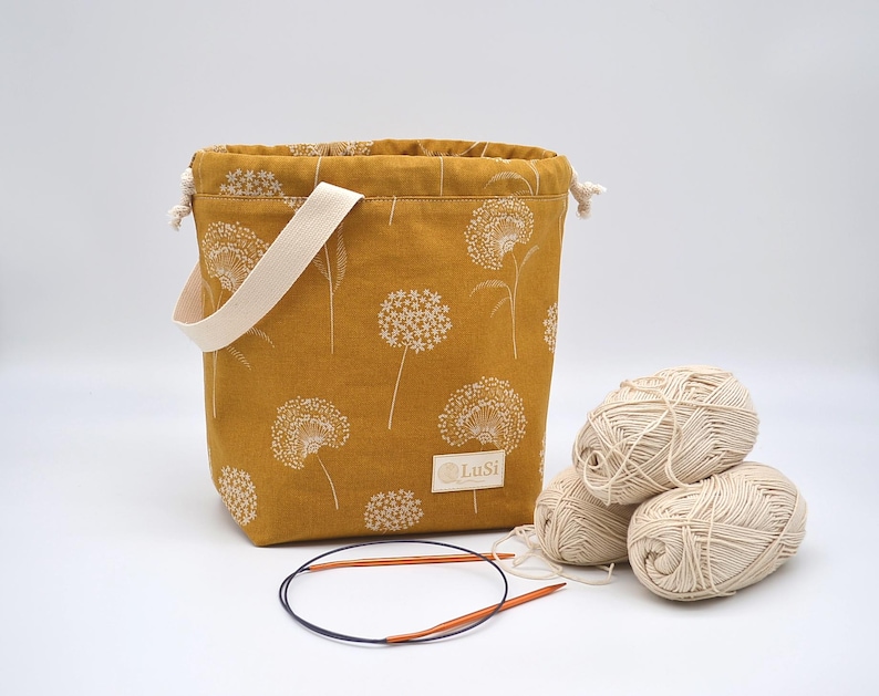 May include: Mustard yellow knitting project bag with a white dandelion pattern, a cream handle, and a drawstring closure. Three balls of cream yarn and a pair of circular knitting needles are next to the bag. The bag has a small label that says "LuSi".