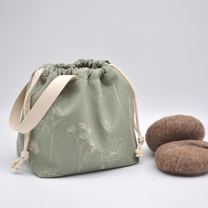 May include: Sage green drawstring bag with a cream-colored handle and floral design. Two brown yarn balls sit beside the bag. The bag is approximately 8 inches wide and 7 inches tall.