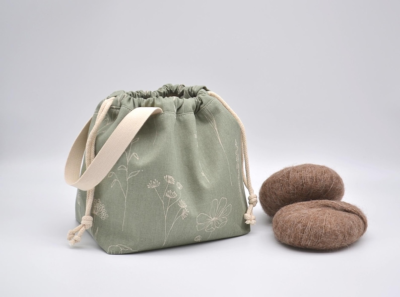 May include: Sage green drawstring bag with a cream-colored handle and floral design. Two brown yarn balls sit beside the bag. The bag is approximately 8 inches wide and 7 inches tall.