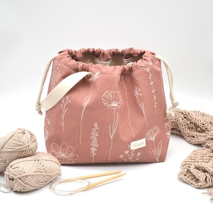 Pode incluir: Uma bolsa de fio com cordão rosa empoeirado com um padrão floral branco, uma alça creme e um cordão creme. Duas bolas de fio bege, agulhas de tricô e um cachecol de malha estão ao lado da bolsa.