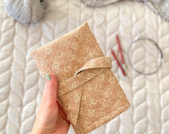 May include: A rectangular fabric case in a tan color with a repeating white geometric pattern. The case features a wrap-around tie closure made of the same material. The exterior has a textured appearance, and the edges are stitched with matching thread.