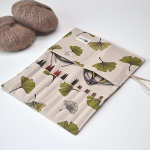 May include: A beige fabric knitting needle case featuring a green and black ginkgo leaf pattern, containing multiple circular knitting needles in various sizes. Two round balls of brown yarn are positioned next to the open needle organiser.