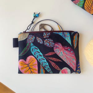 Peut inclure: Une pochette noire zippée ornée d'un motif de feuilles botaniques colorées en rose, bleu et jaune. La pochette est dotée d'une petite poignée noire et d'une boucle en tissu noir. Des ciseaux et une aiguille à tricoter sont visibles à l'intérieur.