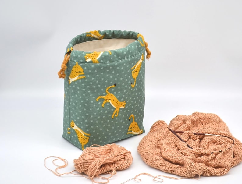 Op de afbeelding: Een groene stoffen projecttas met een patroon van gele cheeta's. De tas is open en toont een cr&egrave;mekleurige binnenkant. Een bol perzikkleurig garen en een gebreide perzikkleurige sjaal met breinaalden liggen naast de tas.