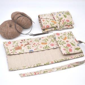May include: A beige and floral patterned knitting needle roll with a small pocket and a brown yarn ball. The roll has a label that says "Lusi" and a brown rabbit illustration.