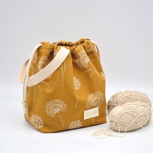 May include: Mustard yellow drawstring bag with a white floral pattern and cream-coloured handles. Two balls of beige yarn sit next to the bag. The bag has a small, rectangular label with the text "LuSi".