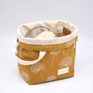 May include: Mustard yellow fabric tote bag with a white dandelion pattern. The bag has a cream-coloured interior and handles. Inside the bag are knitting supplies, including yarn and needles. The label reads "LuSi".